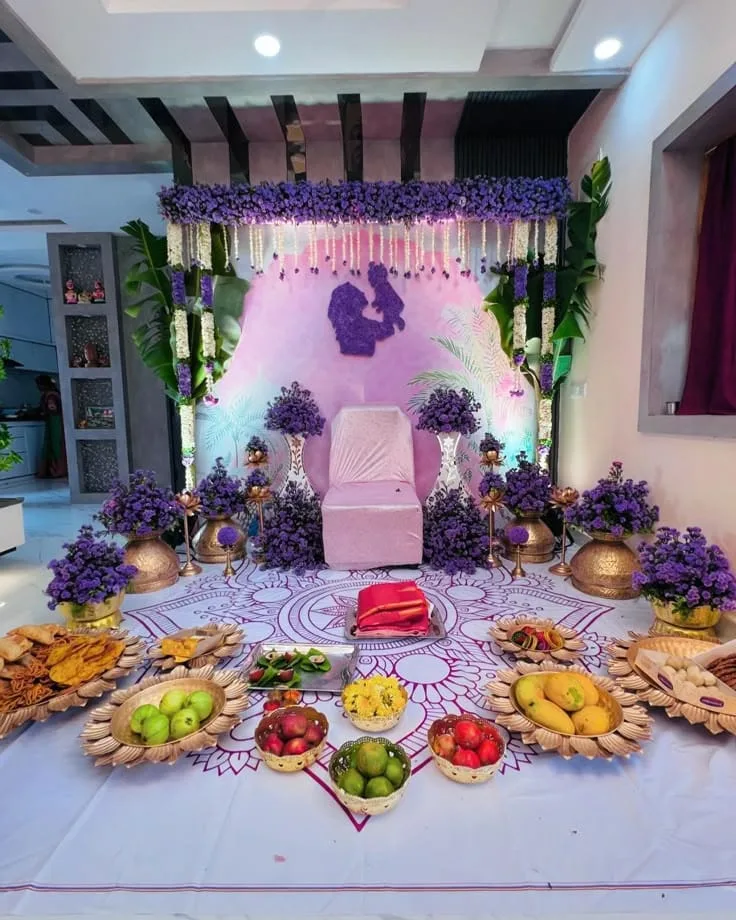 Pink Sreemantham backdrop with a purple mother-and-baby silhouette, hanging white flower strings, banana leaves, a central chair and purple florals in brass vessels