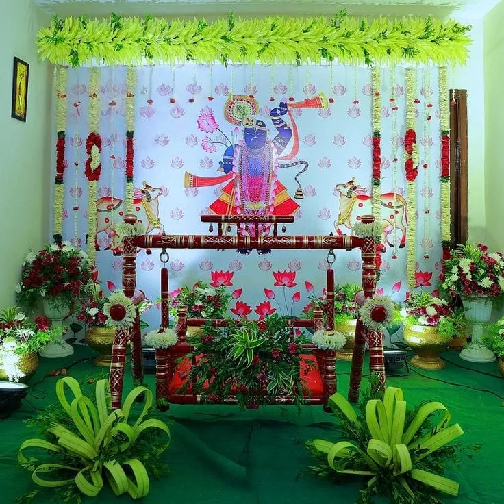 Wooden Barasala cradle before Krishna artwork, surrounded by a leafy canopy, red and white flower strings and green floral arrangements