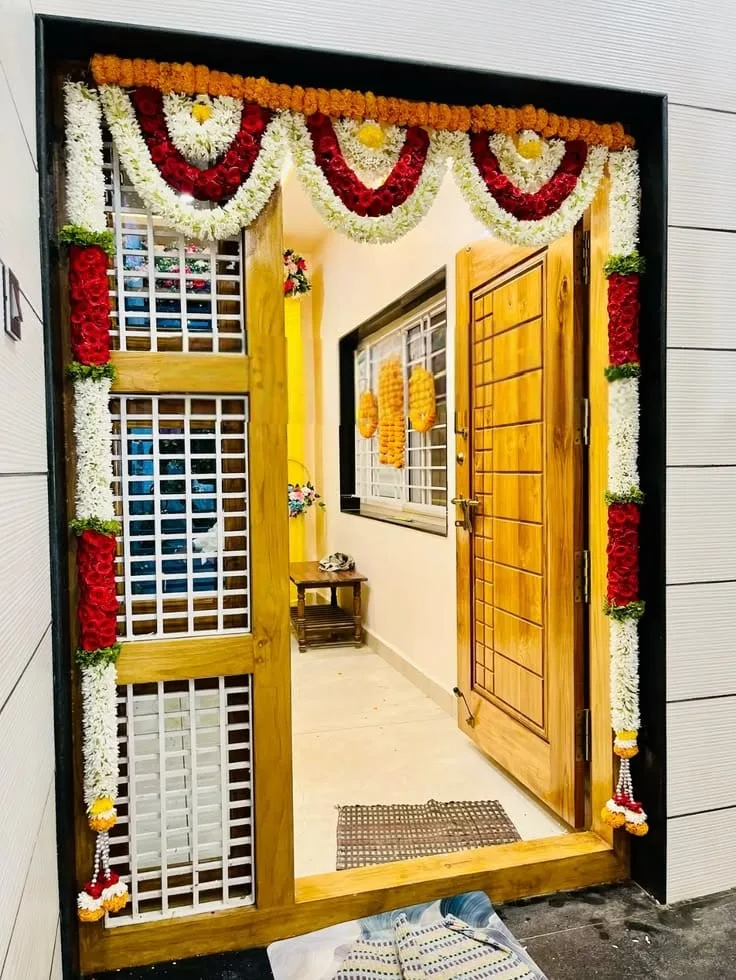Gruhapravesam entrance reference with red and white floral swags, orange flowers across the top and long garlands framing a wooden doorway