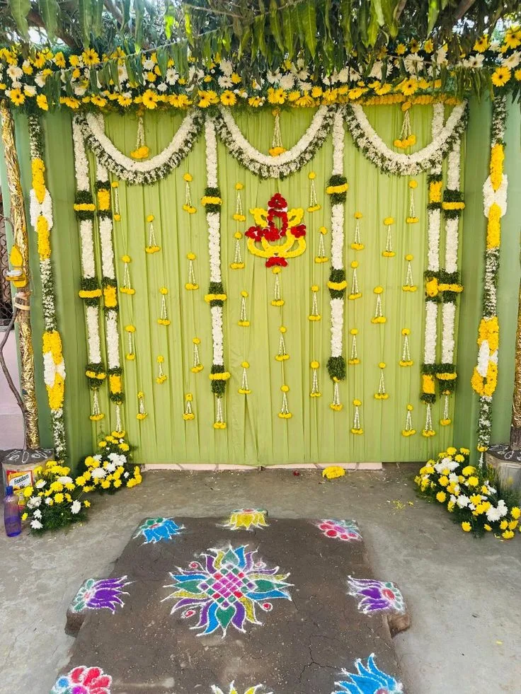 Green Pellikoduku ceremony backdrop with white and yellow floral swags, a red and yellow Ganesh motif, leafy canopy and colourful floor rangoli