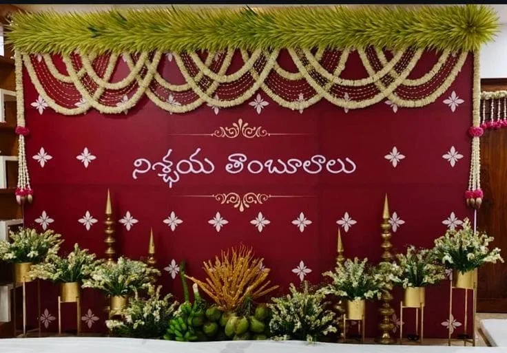 Burgundy Nischaya Tambulam engagement backdrop with Telugu lettering, white flower swags, a leafy canopy, brass lamps and floral stands