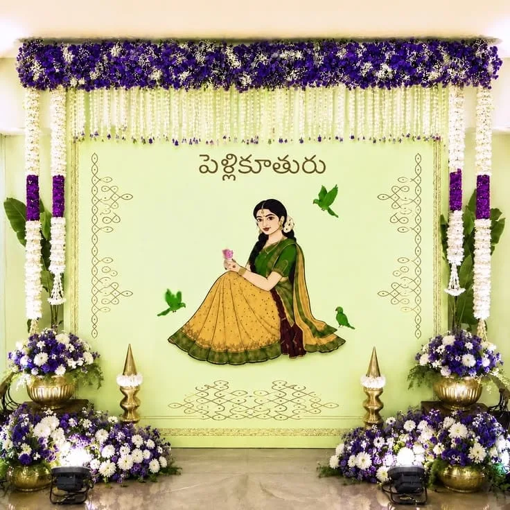 Pale green Telugu Pellikuturu backdrop with an illustrated bride, purple and white flower borders, hanging flower strings and brass lamps