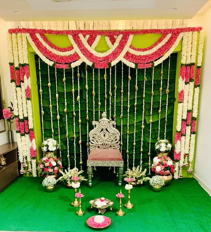 Green half saree stage reference with pink and white flower swags, hanging garlands, an ornate chair and brass lamps